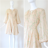 A vintage-style cream or beige dress with a green and yellow floral branch print, displayed on a white mannequin. The image features a full-length view on the left and a close-up detail of the pattern on the right.